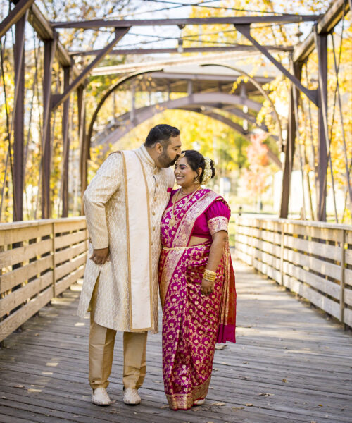 A couple in traditional Indian attire smiles on a wooden bridge surrounded by autumn trees.