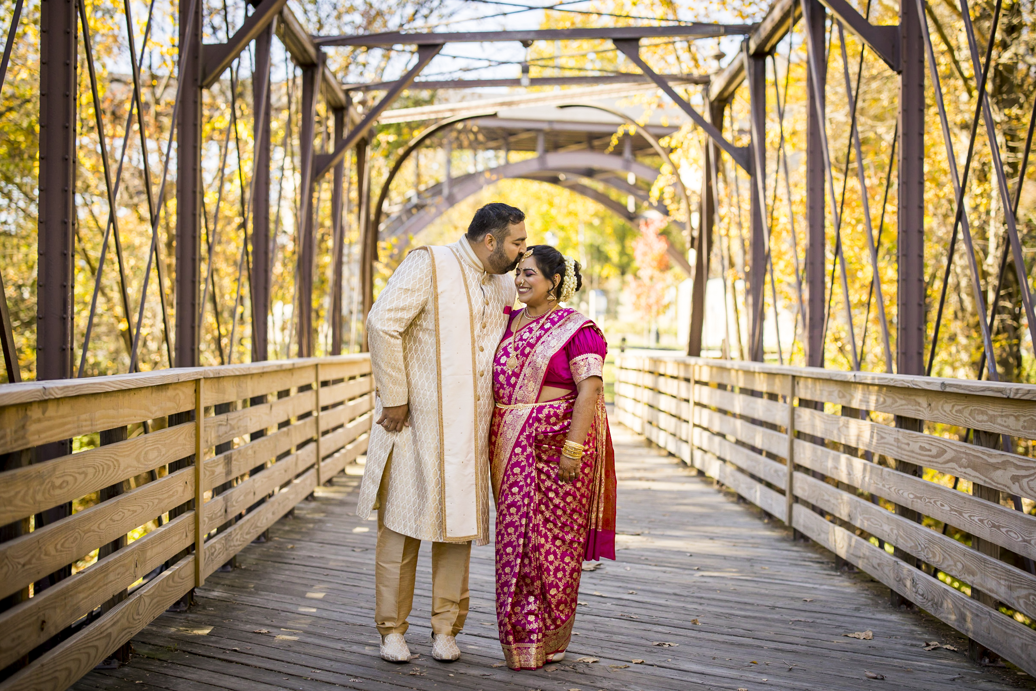 A couple in traditional Indian attire smiles on a wooden bridge surrounded by autumn trees.