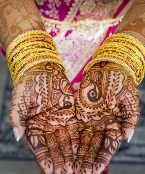 Hands with intricate henna designs, adorned with gold bangles, held together over a pink saree.