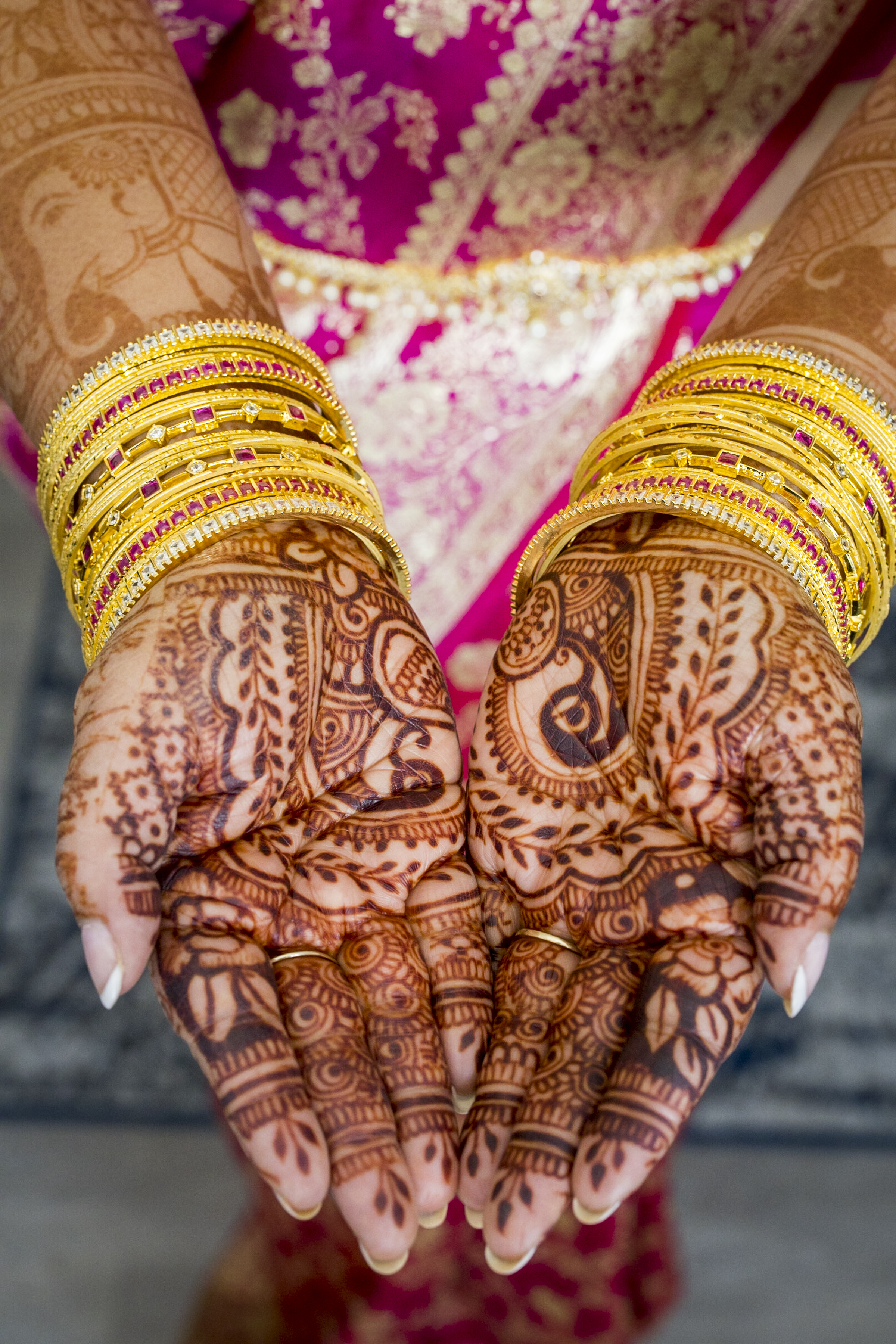 Hands with intricate henna designs, adorned with gold bangles, held together over a pink saree.