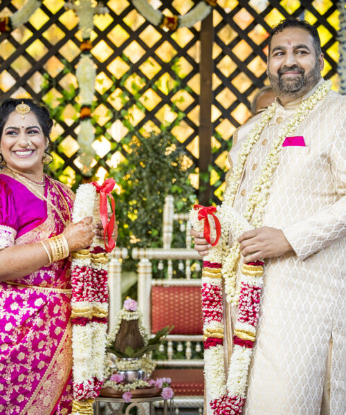 A bride and groom in traditional Indian attire smile, holding flower garlands at their wedding ceremony.