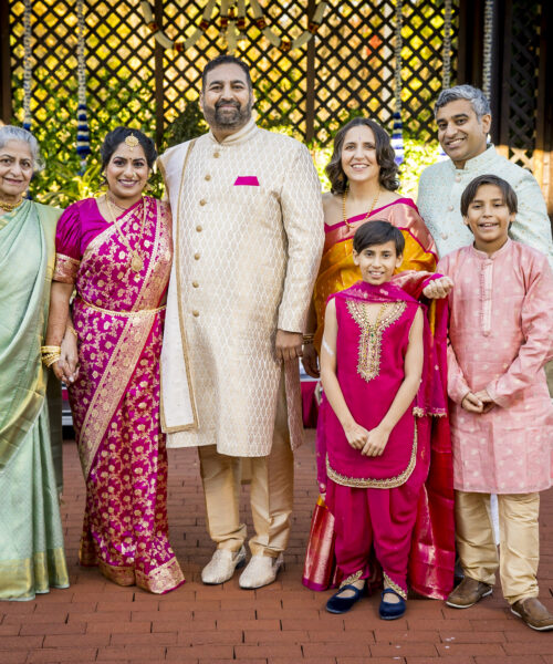 A family in colorful traditional Indian attire poses and smiles outdoors in front of a wooden lattice.