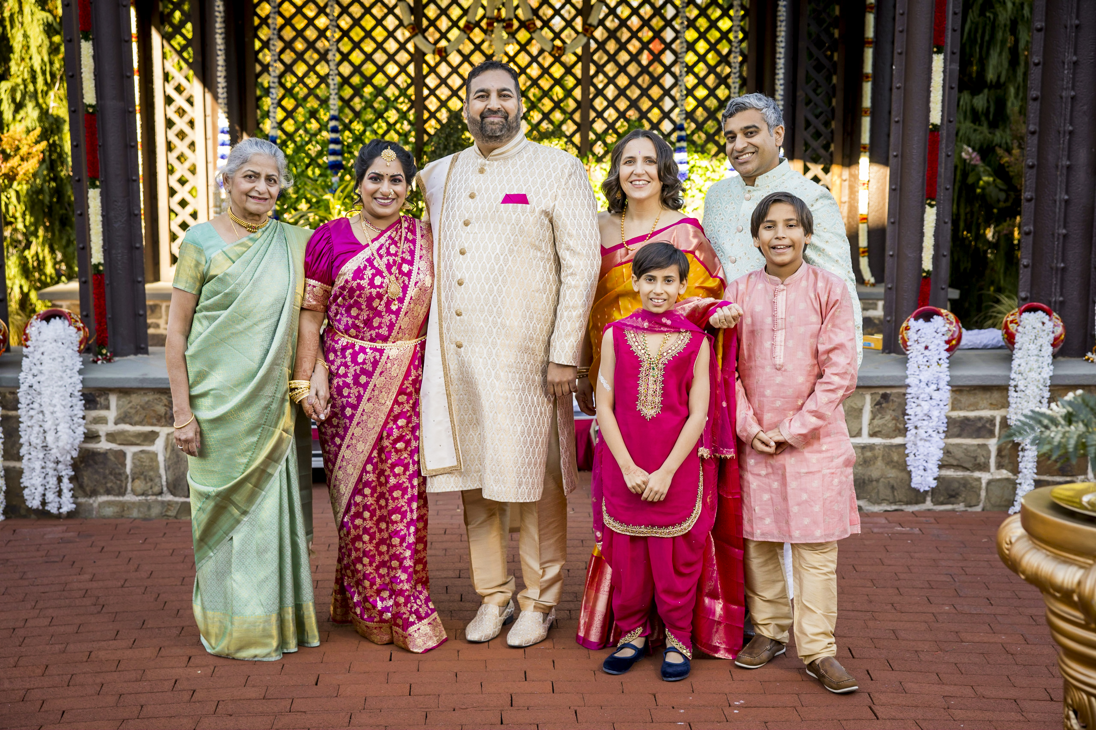 A family in colorful traditional Indian attire poses and smiles outdoors in front of a wooden lattice.