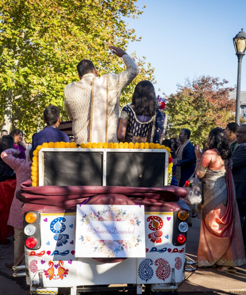 A couple in traditional attire rides in a decorated vehicle, surrounded by a festive crowd outdoors.