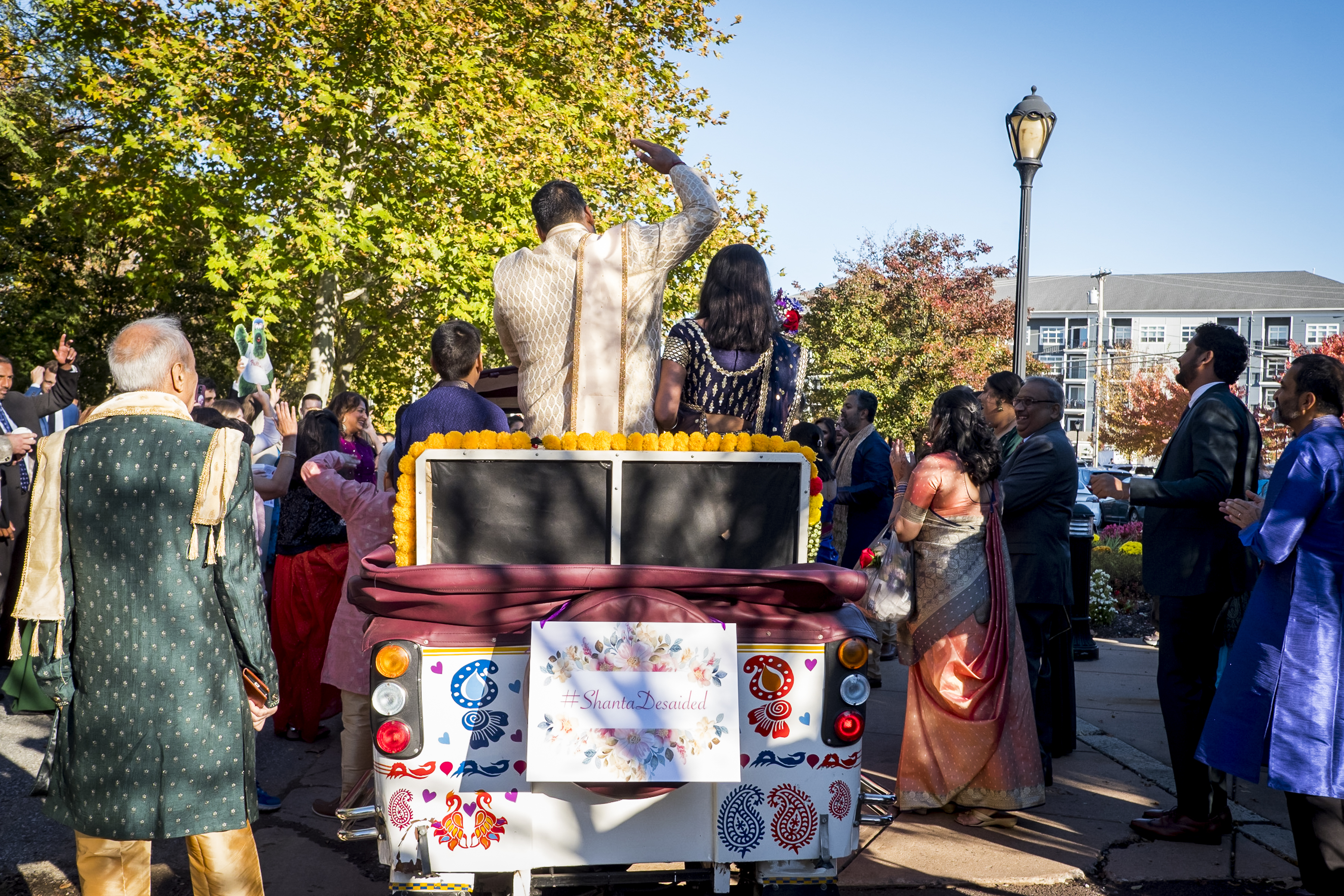 A couple in traditional attire rides in a decorated vehicle, surrounded by a festive crowd outdoors.