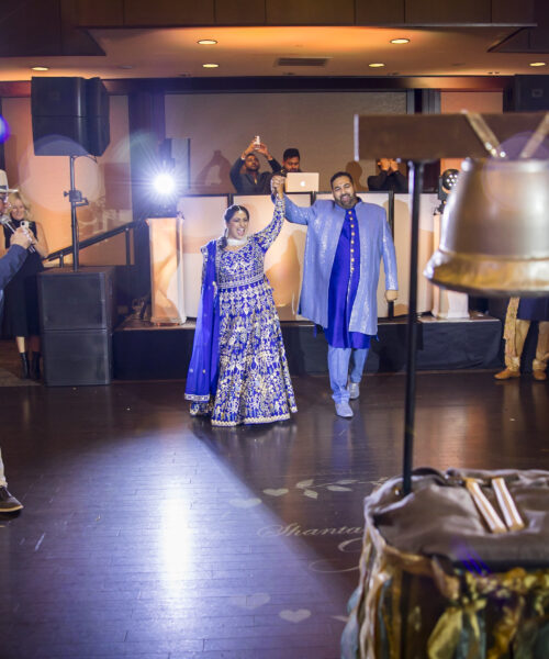 A couple in blue traditional attire stands on a dance floor, surrounded by people clapping and a large silver bell.