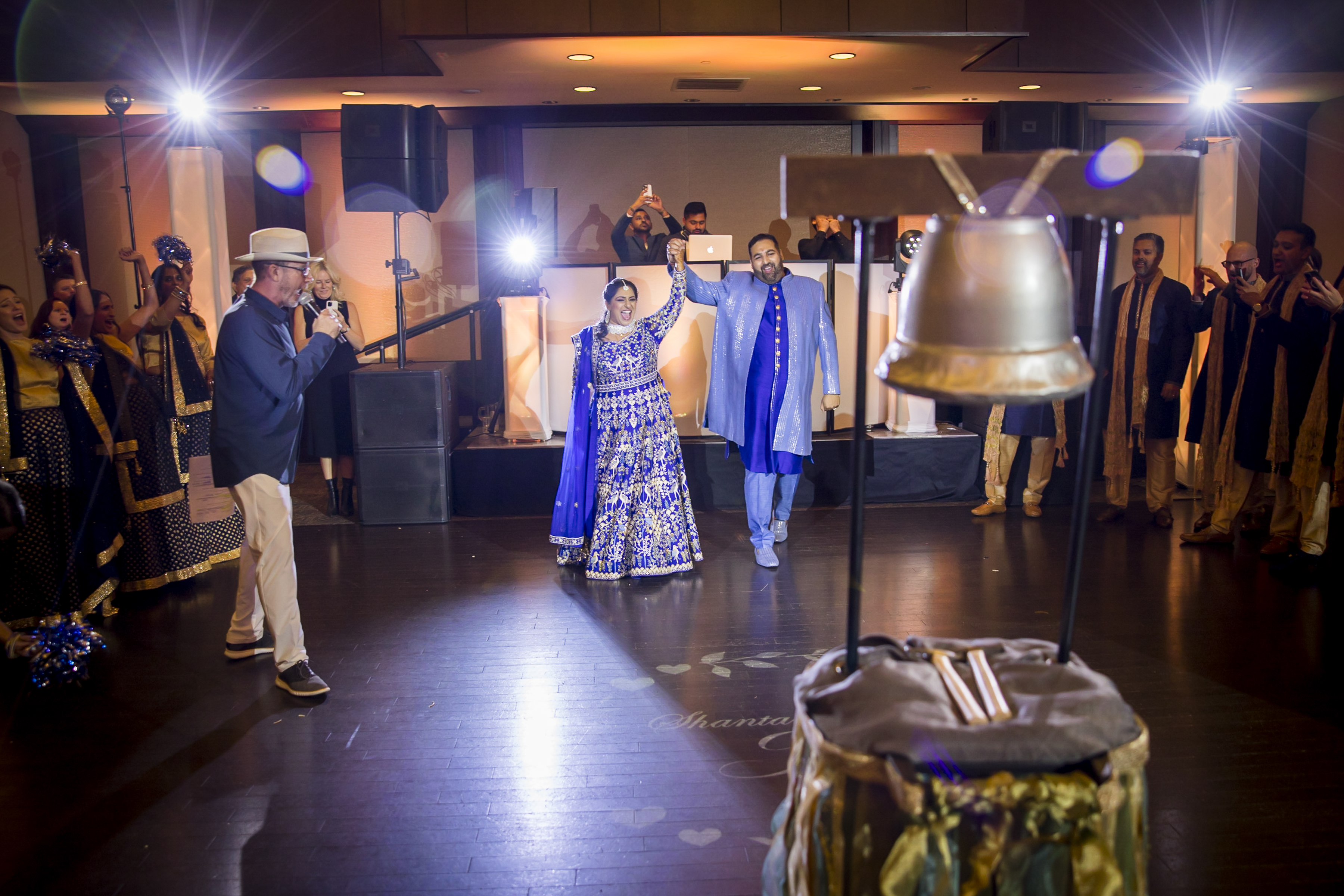A couple in blue traditional attire stands on a dance floor, surrounded by people clapping and a large silver bell.