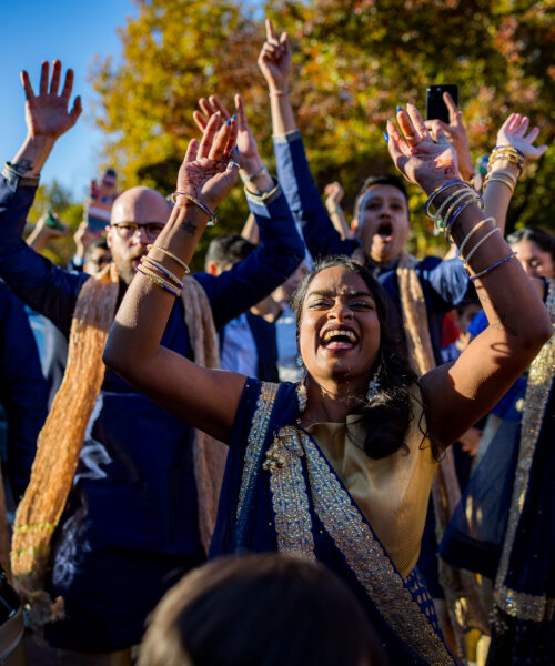A joyful group in festive attire celebrates outdoors, raising their hands and smiling under a bright sky.