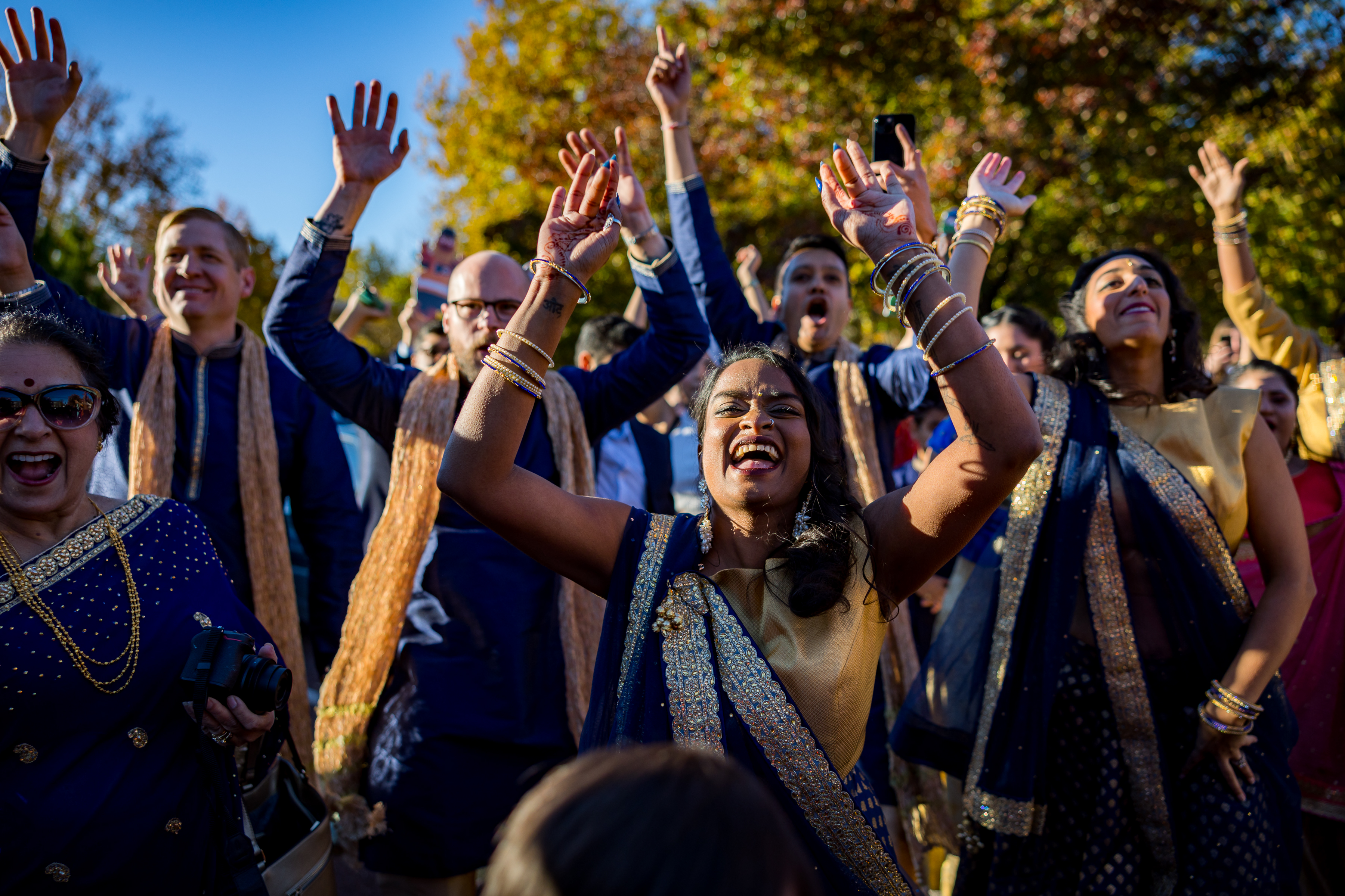A joyful group in festive attire celebrates outdoors, raising their hands and smiling under a bright sky.