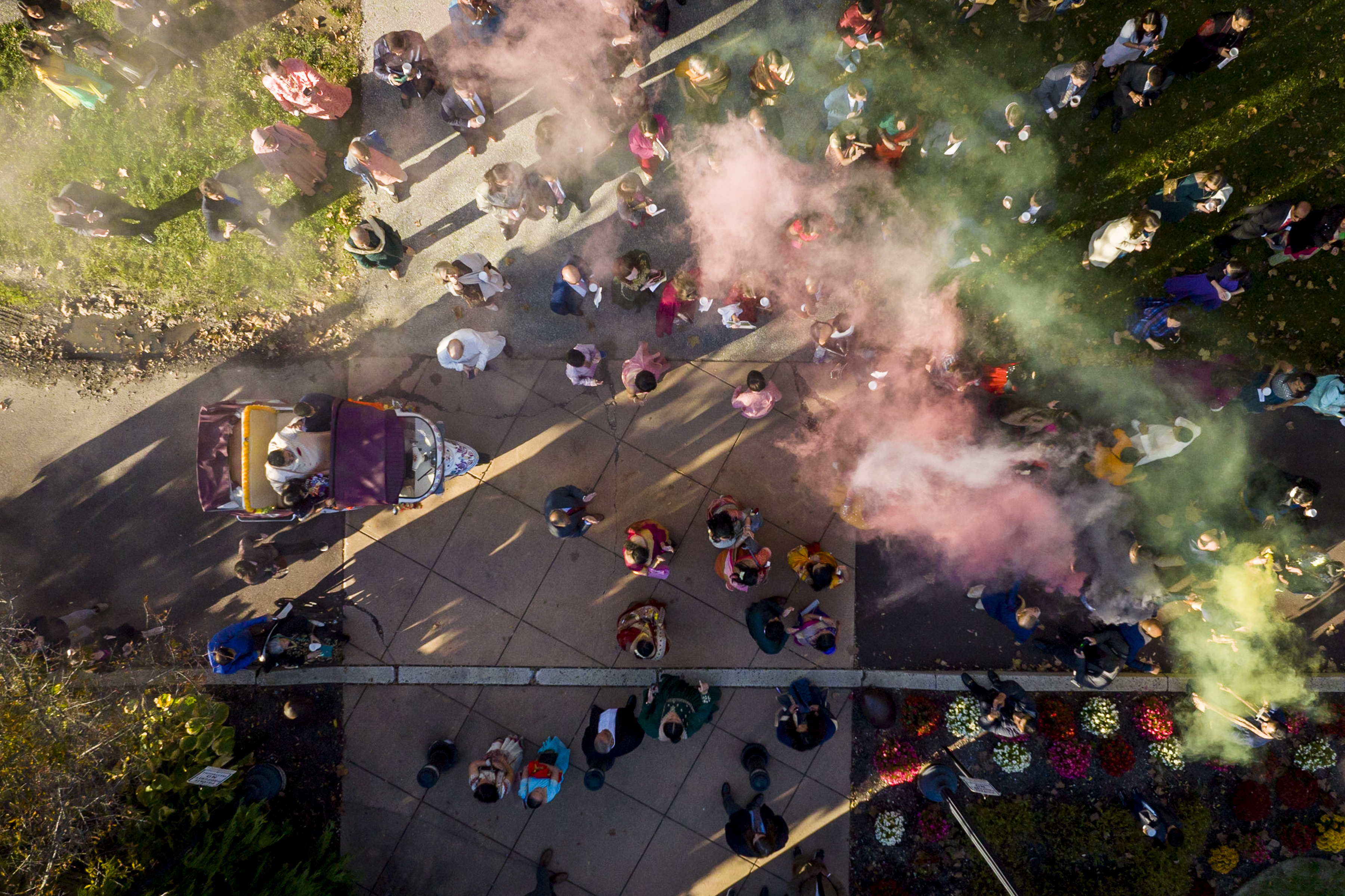 Aerial view of a crowd on a path with colorful smoke and sunlight, surrounded by greenery and flowers.
