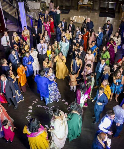 A large, colorful crowd dances at a lively indoor celebration, viewed from above.