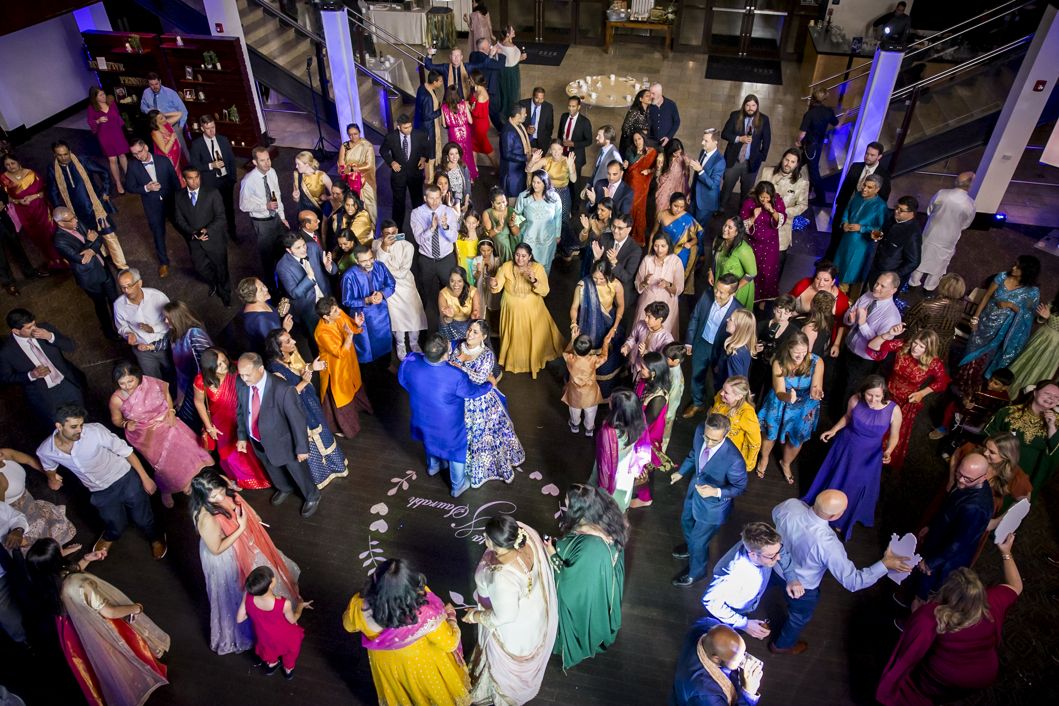 A large, colorful crowd dances at a lively indoor celebration, viewed from above.