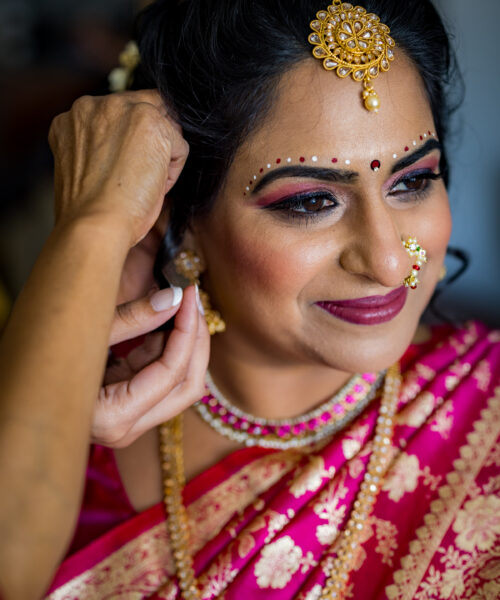 A woman in a pink saree and gold jewelry smiles as someone helps her put on an earring.