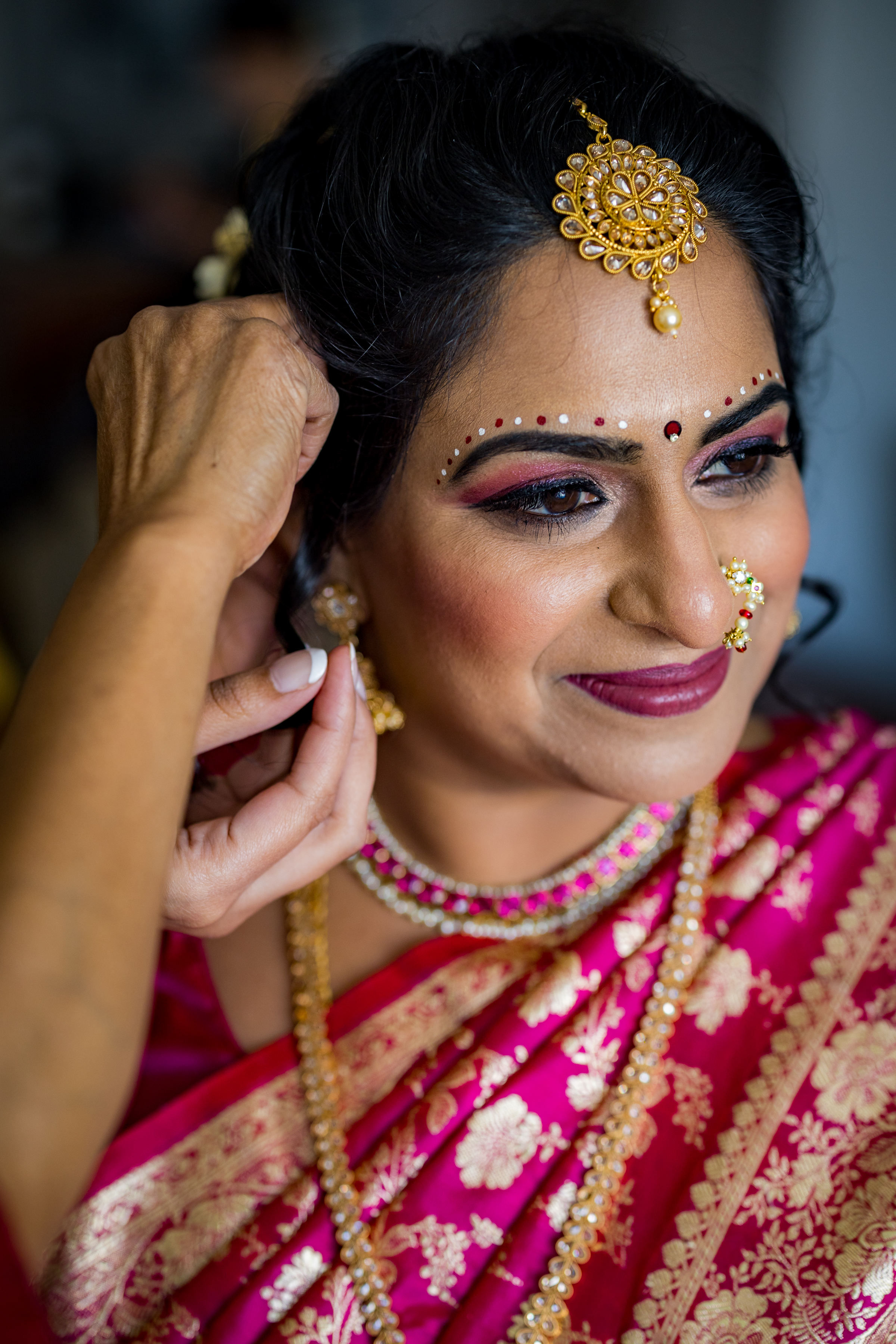 A woman in a pink saree and gold jewelry smiles as someone helps her put on an earring.