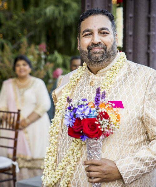 Smiling man in traditional attire holds a bouquet and wears a garland, with a woman and chairs in the background.