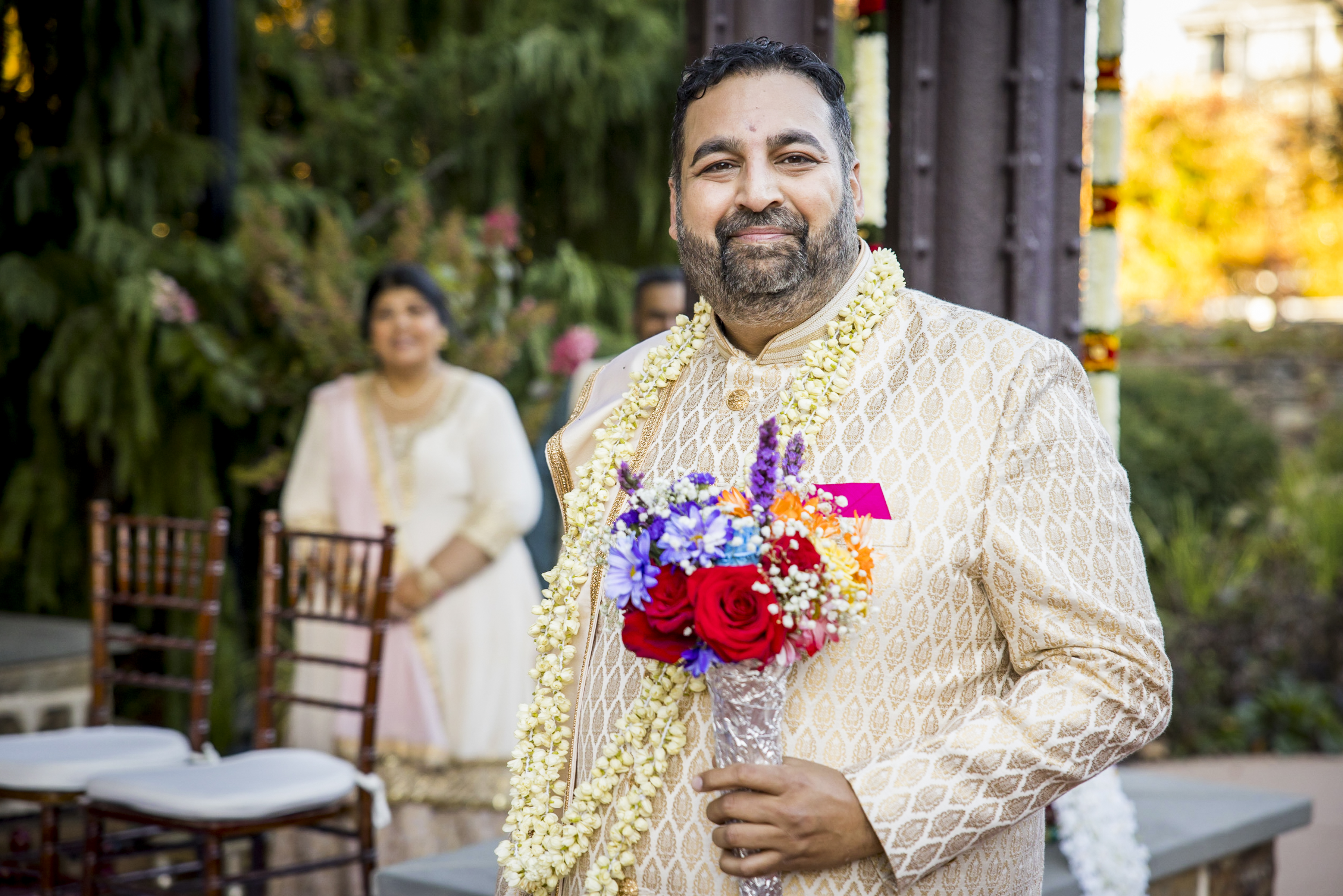Smiling man in traditional attire holds a bouquet and wears a garland, with a woman and chairs in the background.