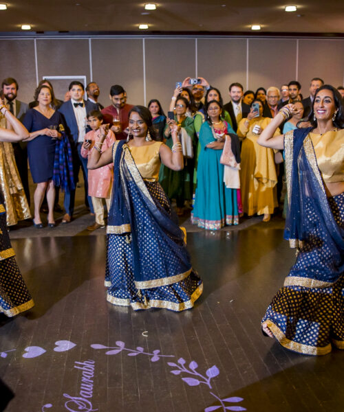 Women in navy and gold traditional outfits dance joyfully at a celebration, while a crowd watches and smiles.