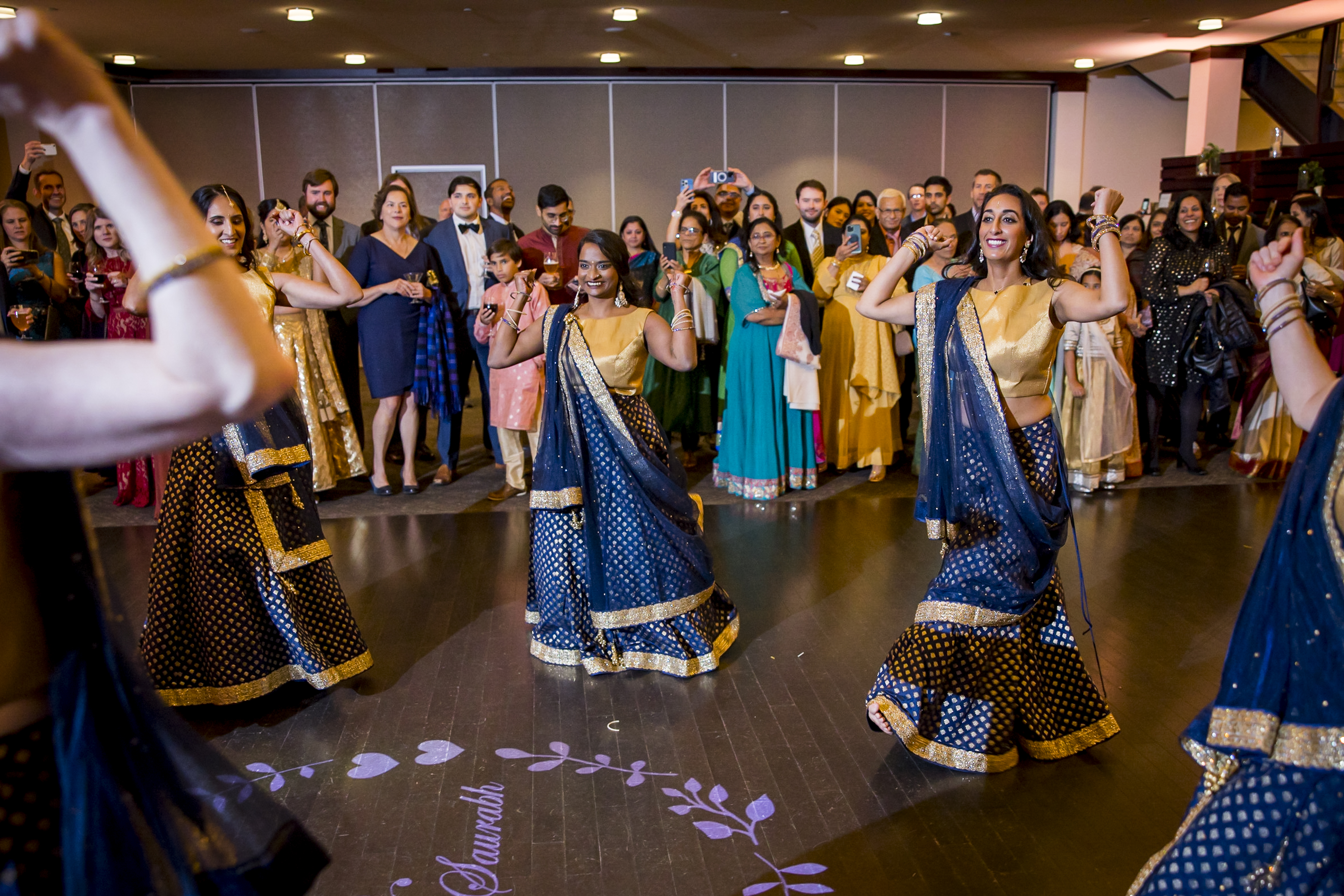 Women in navy and gold traditional outfits dance joyfully at a celebration, while a crowd watches and smiles.