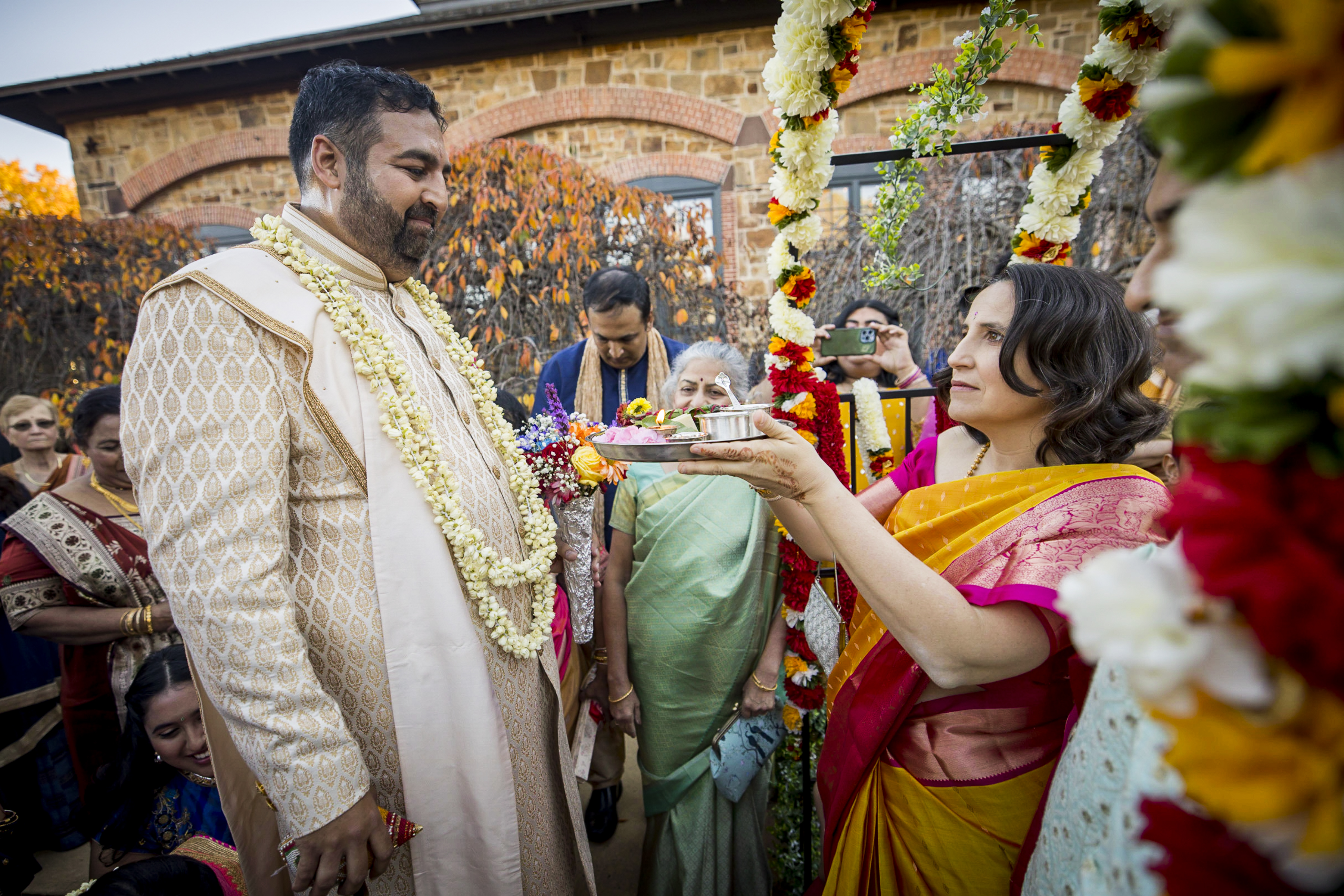 A woman in a sari performs a ritual for a man in traditional attire at an outdoor Indian wedding ceremony.