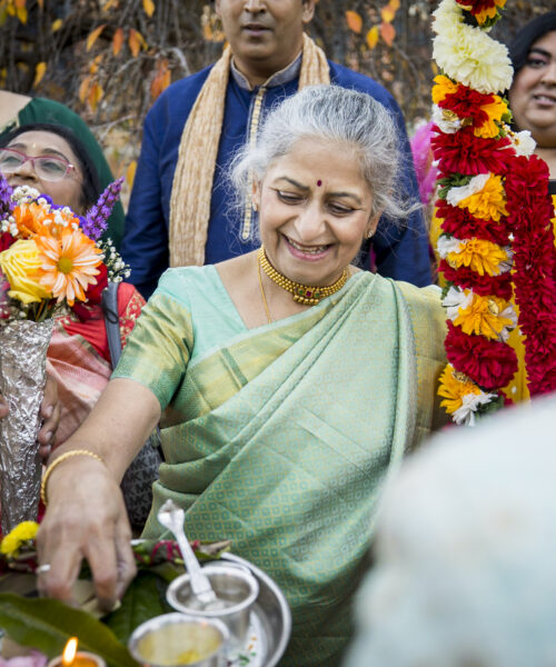 An elderly woman in a green sari smiles while holding a ceremonial tray at a festive outdoor gathering.