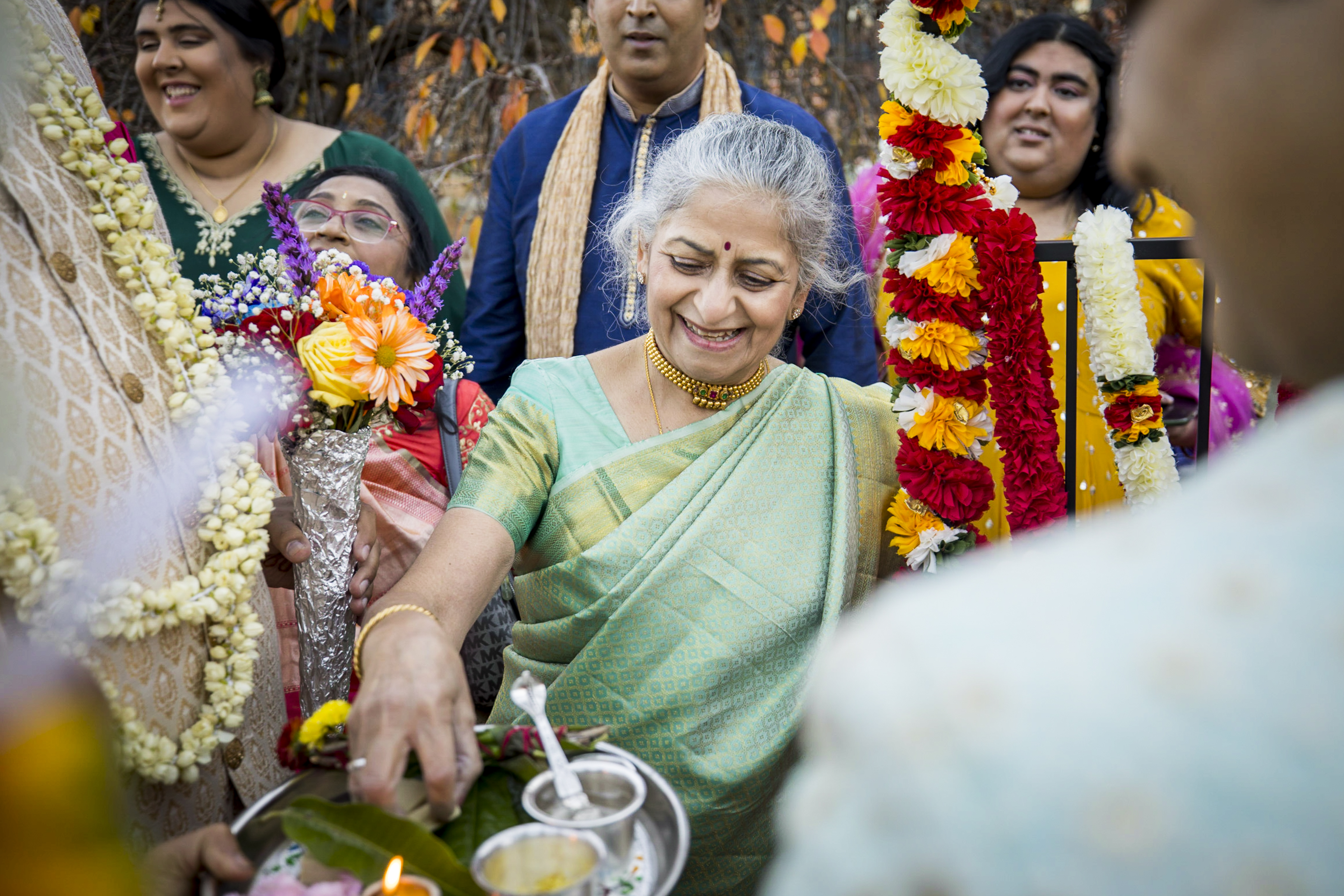 An elderly woman in a green sari smiles while holding a ceremonial tray at a festive outdoor gathering.