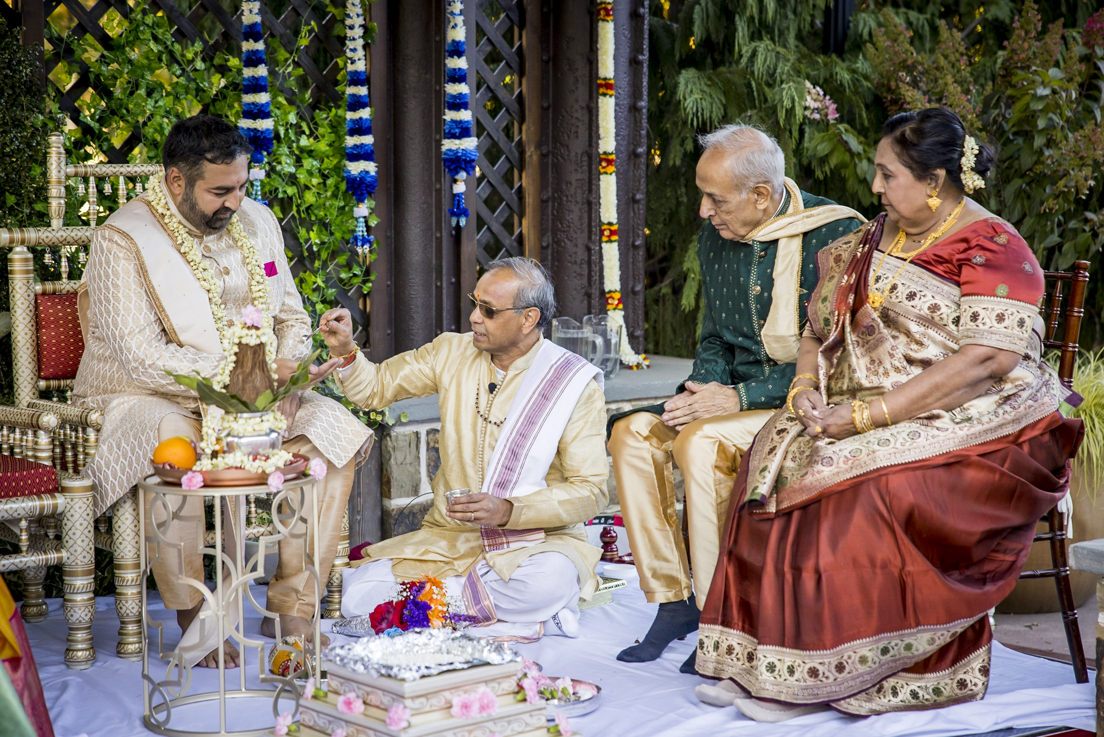 A Hindu wedding ceremony with four adults in traditional attire performing rituals outdoors.