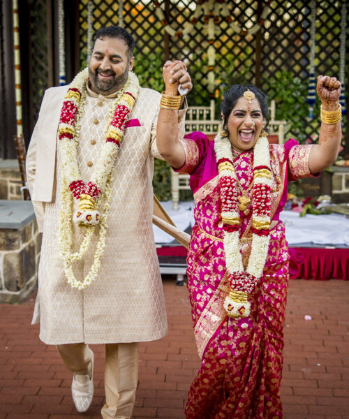 A joyful couple in traditional Indian wedding attire hold hands and celebrate outdoors.
