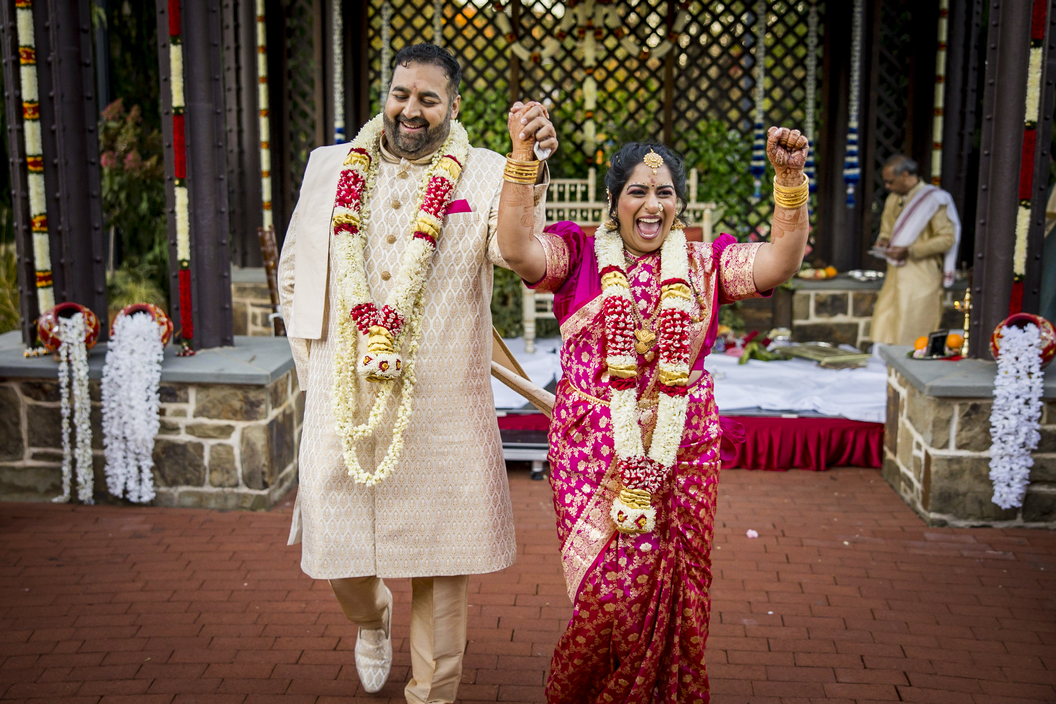 A joyful couple in traditional Indian wedding attire hold hands and celebrate outdoors.