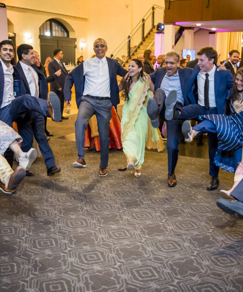 A group of people in formal and traditional attire dance joyfully in a circle, kicking their legs up in unison.
