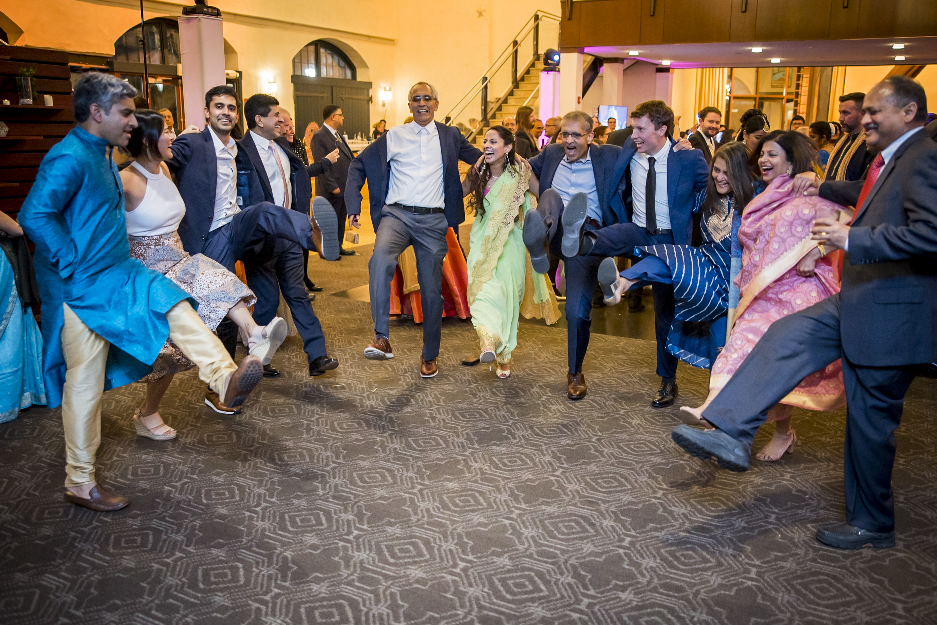 A group of people in formal and traditional attire dance joyfully in a circle, kicking their legs up in unison.
