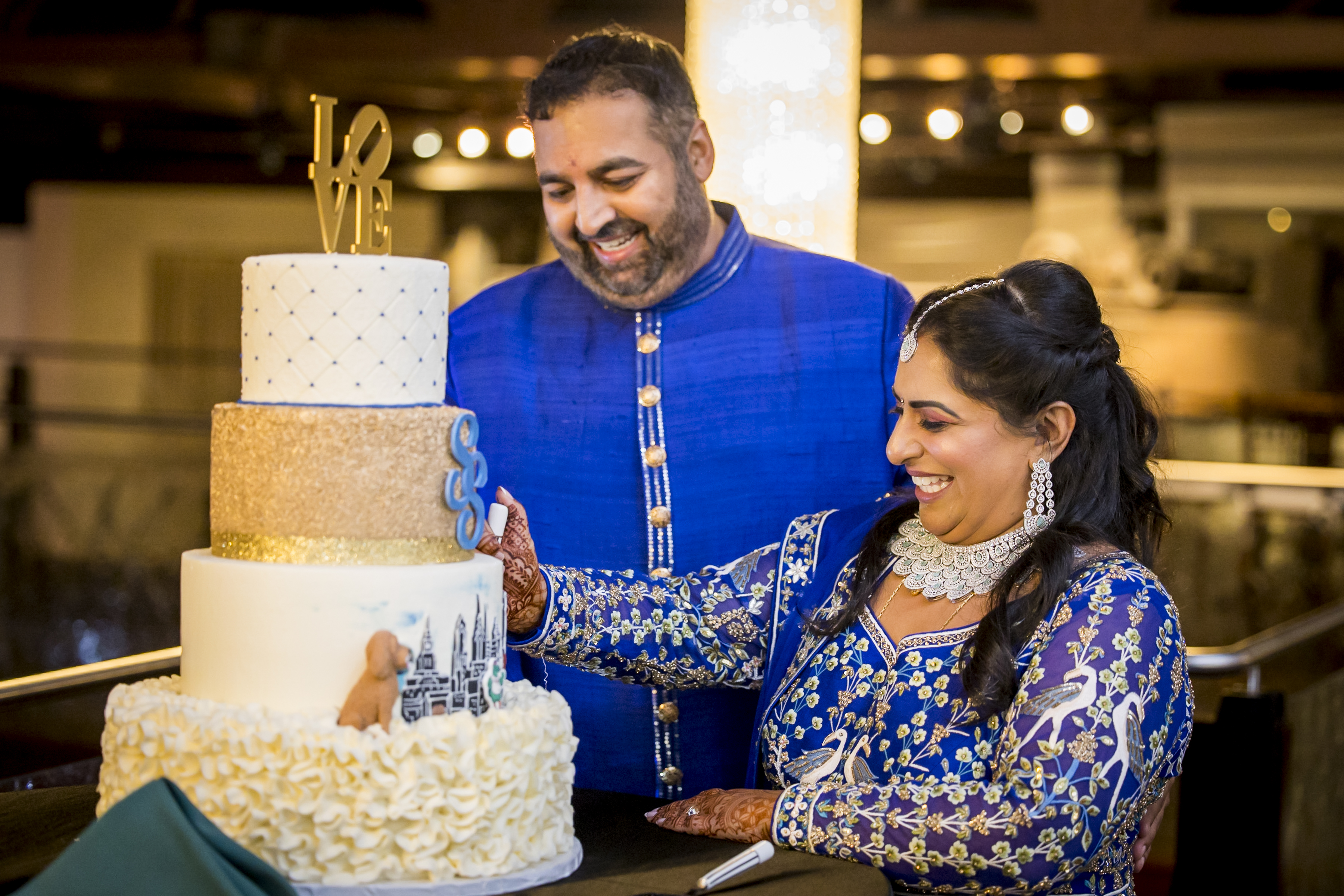 A smiling couple in blue outfits cut a three-tiered cake at a celebration.