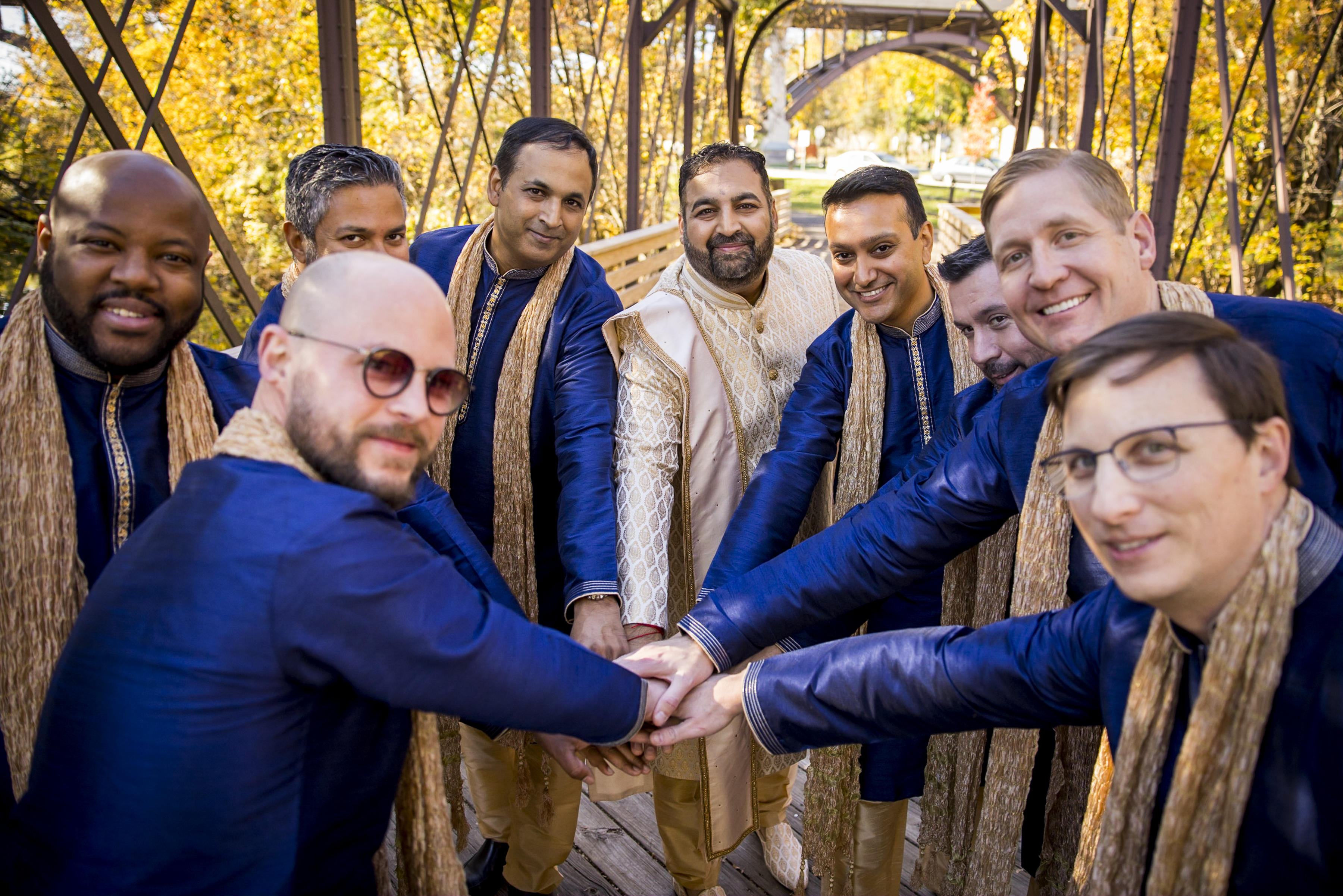 A group of men in matching blue and gold outfits stack hands on a bridge surrounded by fall foliage.