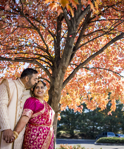 A couple in traditional attire stands under a vibrant autumn tree, holding hands and smiling.