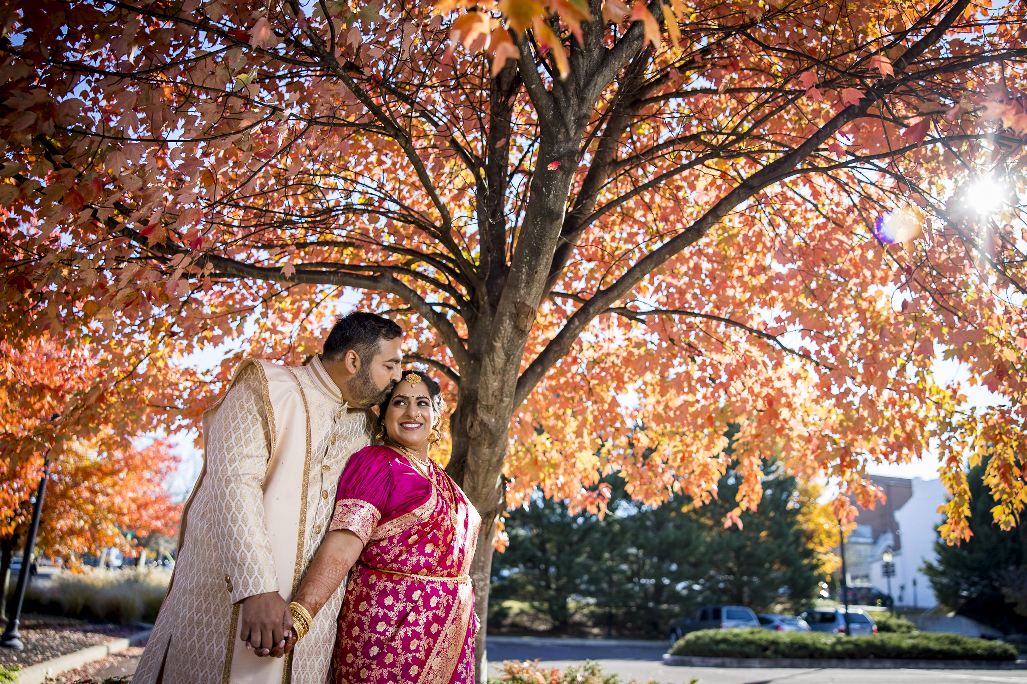 A couple in traditional attire stands under a vibrant autumn tree, holding hands and smiling.