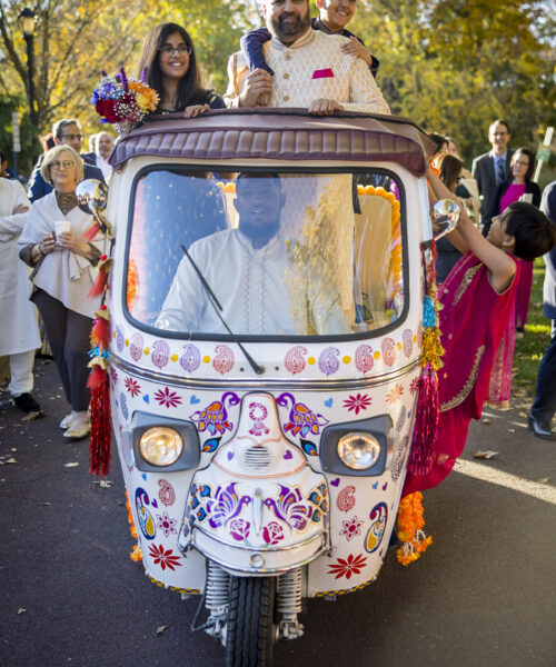 A family in traditional attire rides a decorated tuk-tuk at an outdoor celebration with people in the background.