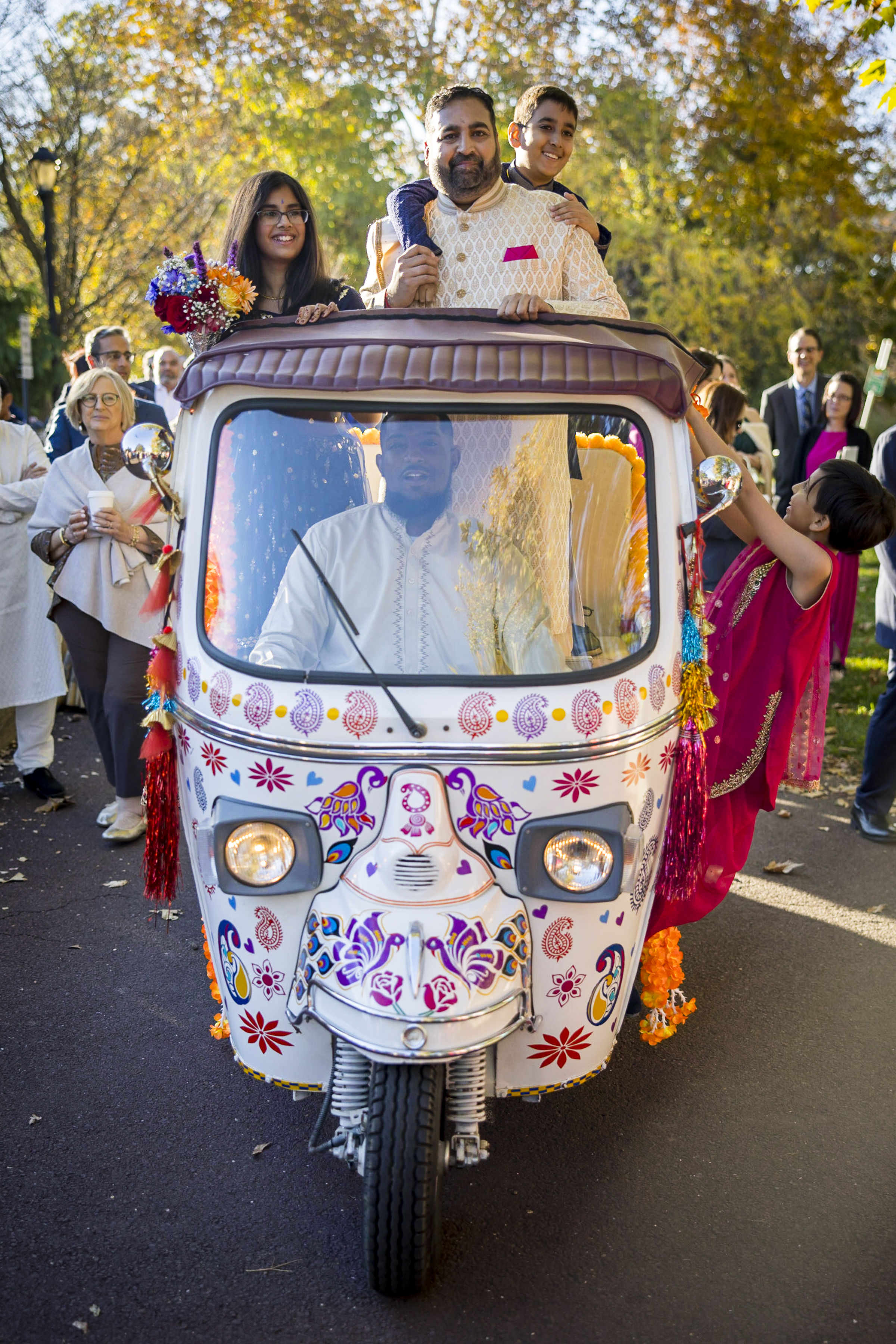 A family in traditional attire rides a decorated tuk-tuk at an outdoor celebration with people in the background.
