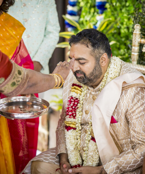 A woman performs a traditional blessing on a seated man at an Indian wedding ceremony.
