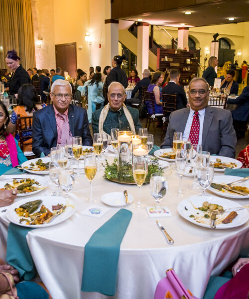 Eight people in formal attire sit around a round table with food and drinks at a festive indoor event.