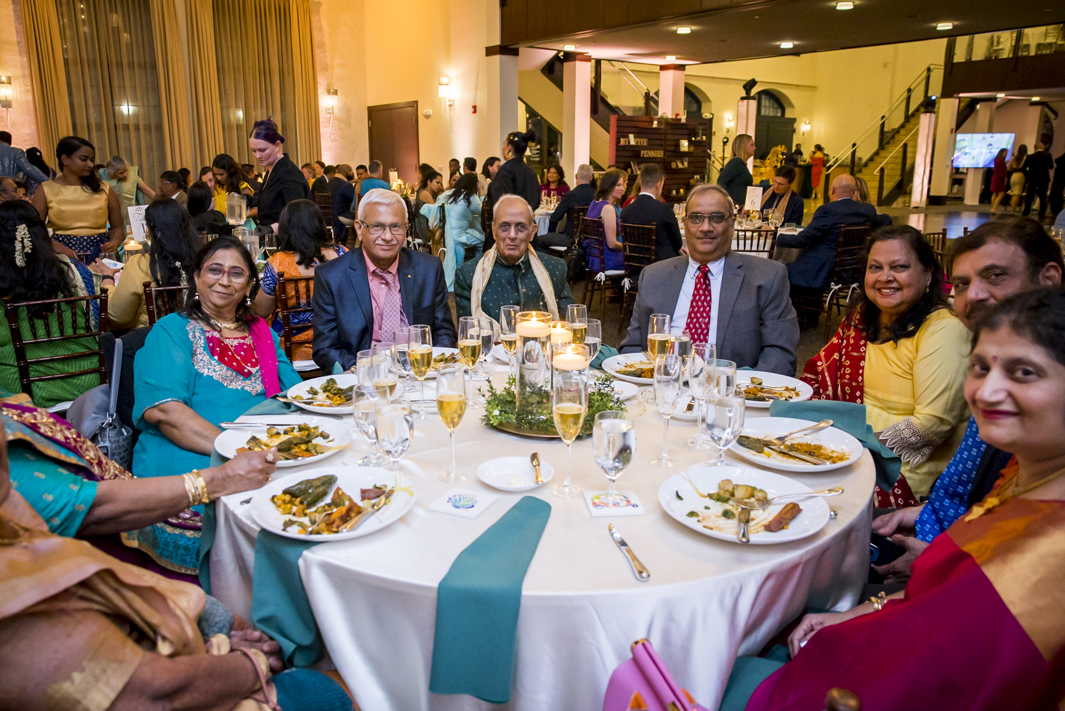 Eight people in formal attire sit around a round table with food and drinks at a festive indoor event.