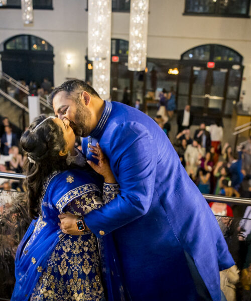 A couple in blue traditional attire kisses on a balcony at a festive indoor event with people below.