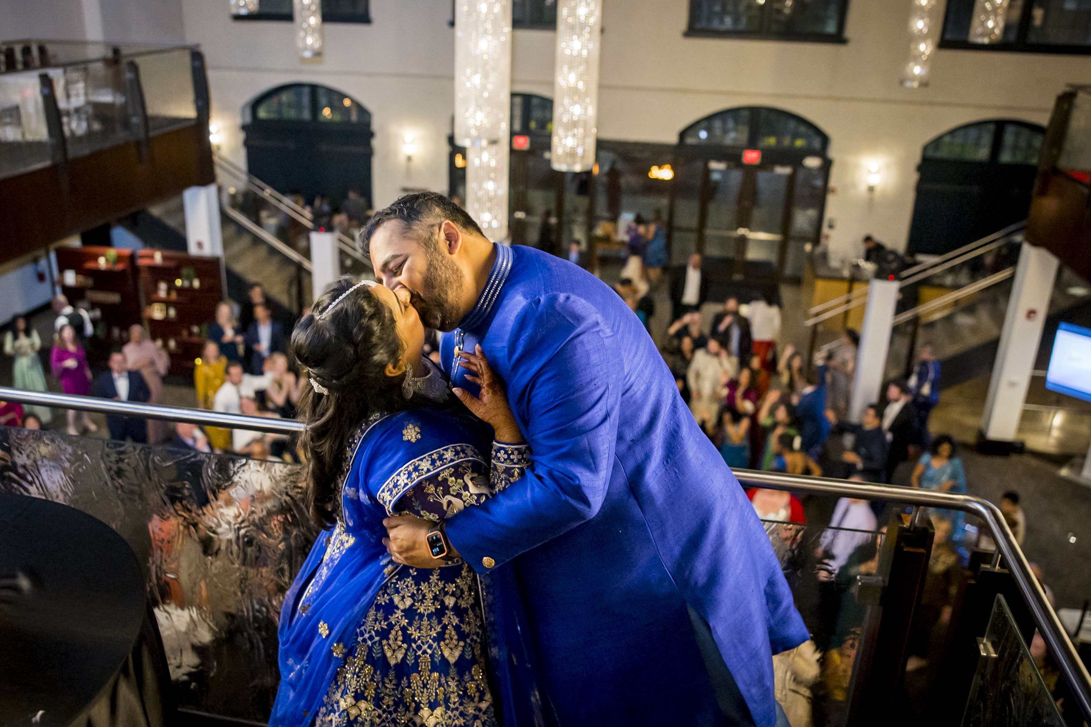 A couple in blue traditional attire kisses on a balcony at a festive indoor event with people below.