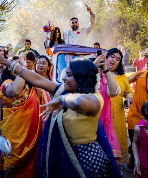 People in colorful traditional clothing joyfully dance outdoors around a car during a festive celebration.