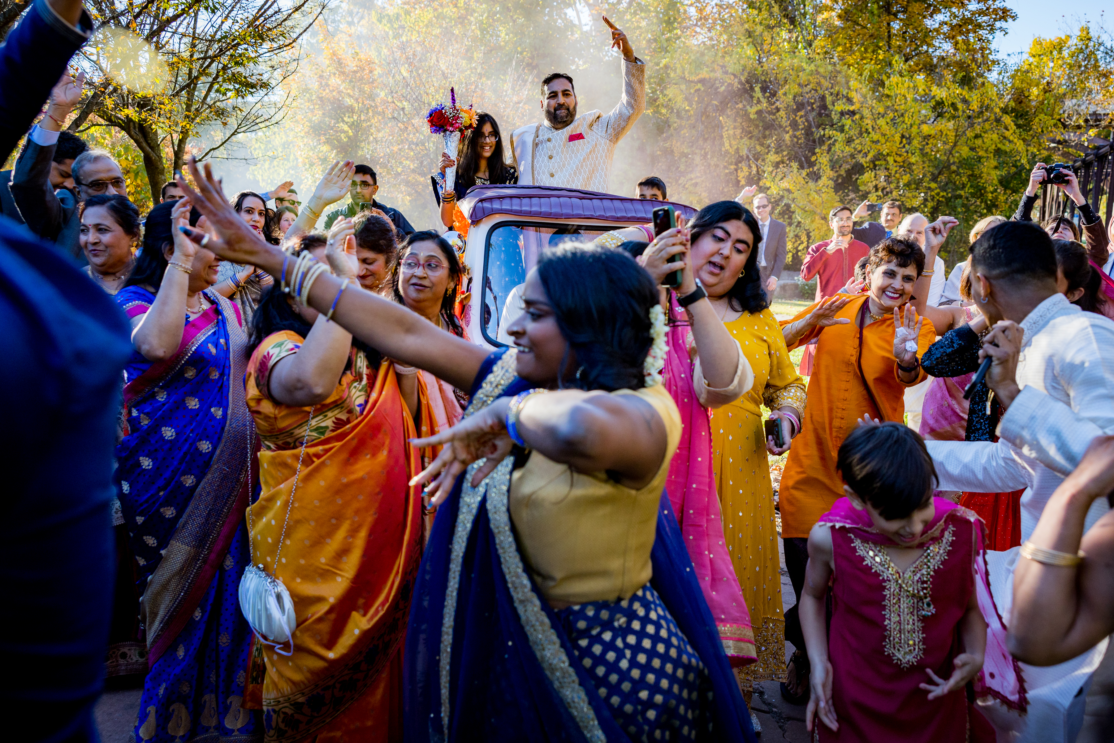 People in colorful traditional clothing joyfully dance outdoors around a car during a festive celebration.
