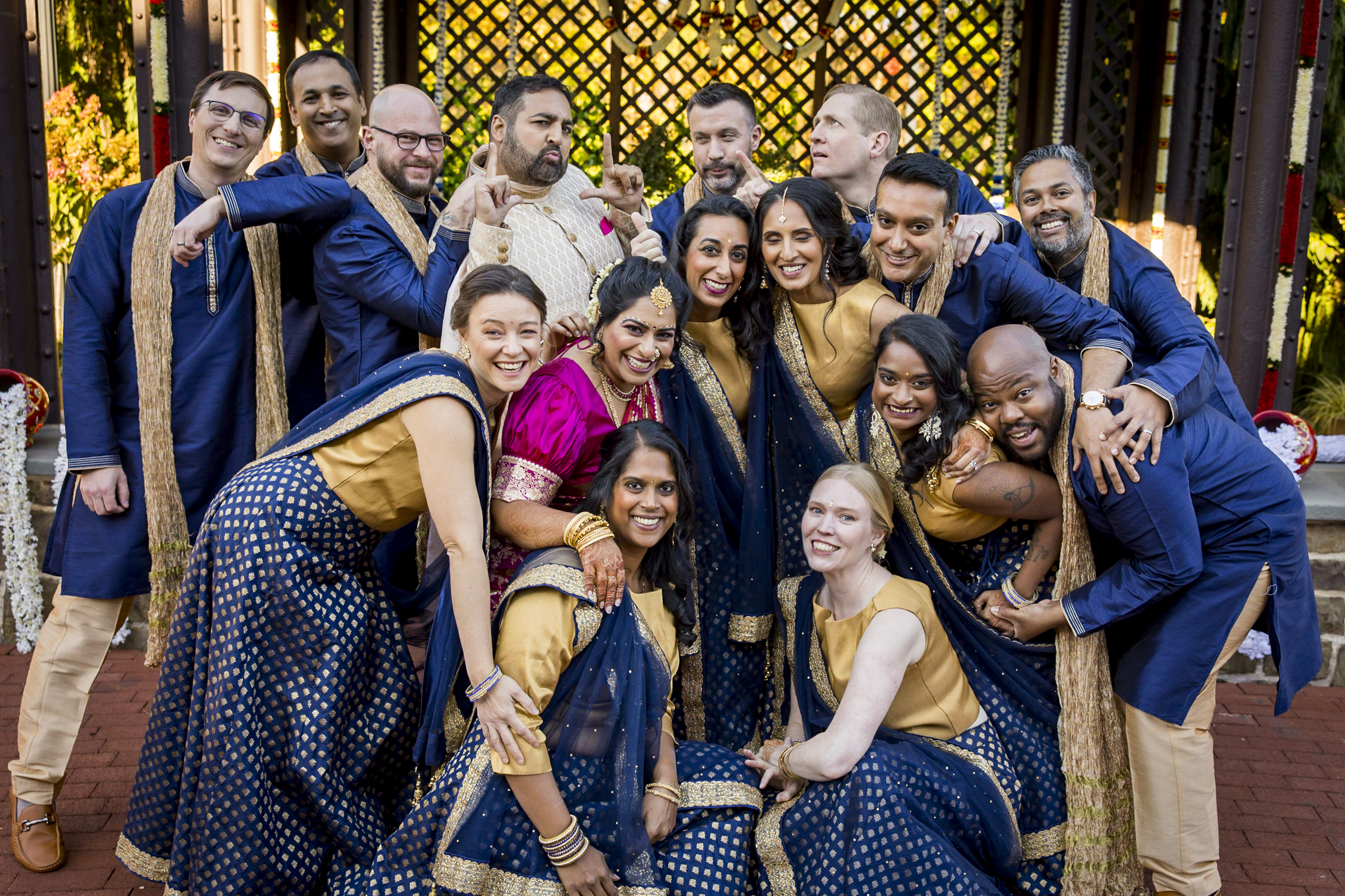 A joyful group in coordinated Indian attire poses together, smiling and laughing outdoors.