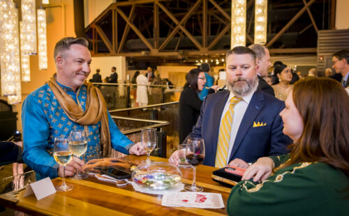 Three people in formal attire chat at a bar with drinks and a bowl on the counter in a warmly lit venue.