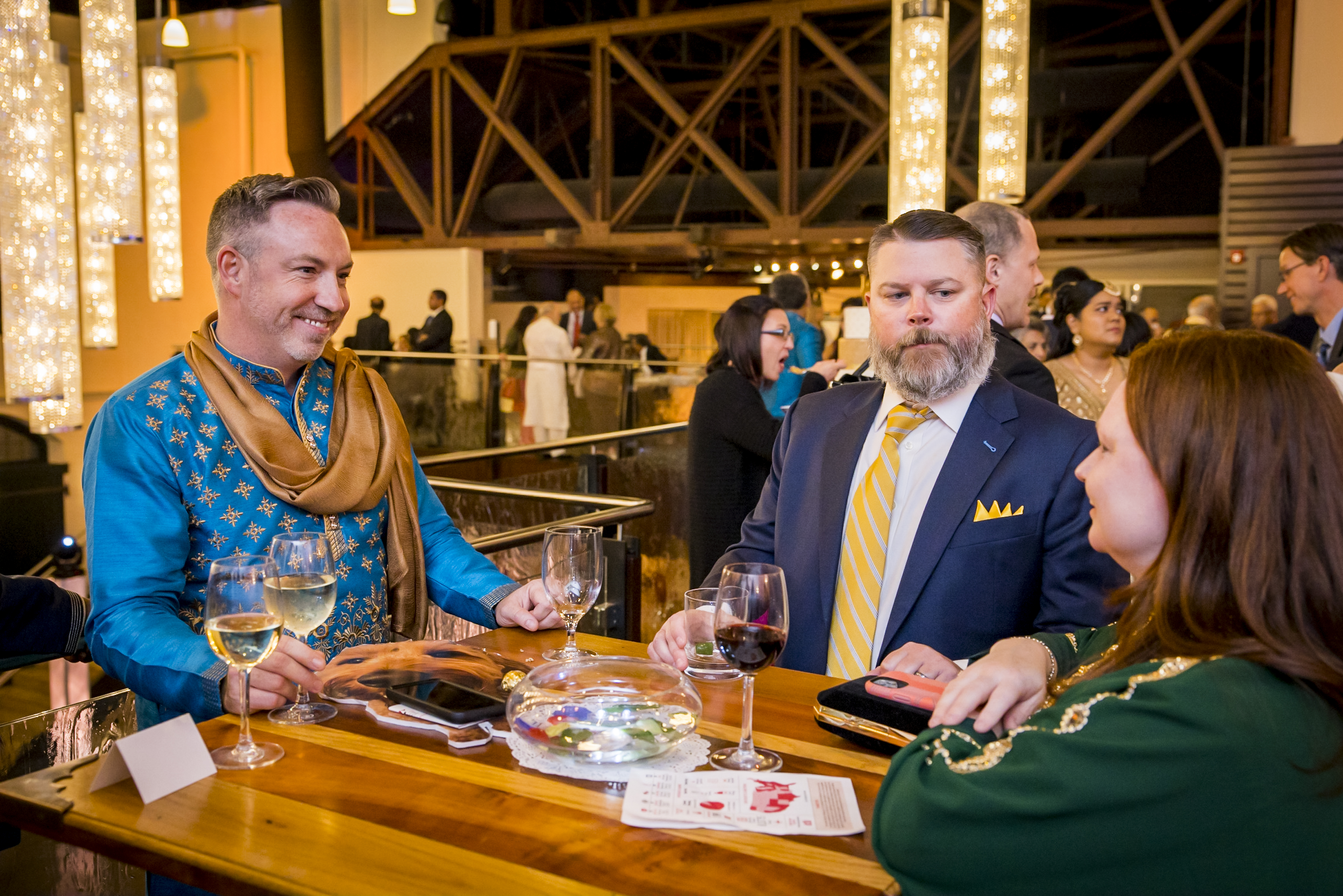 Three people in formal attire chat at a bar with drinks and a bowl on the counter in a warmly lit venue.