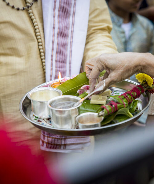 Hands holding a decorated puja thali with flowers, leaves, and diya during an Indian ritual or ceremony.