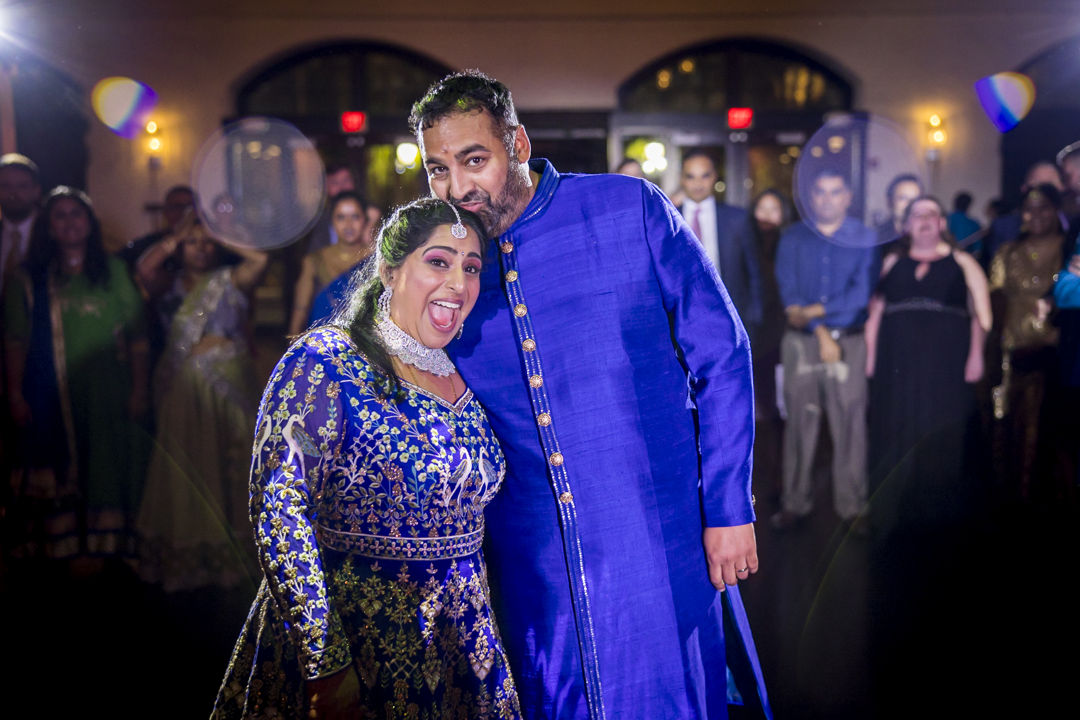 A joyful couple in blue traditional attire pose together, smiling at a festive indoor event with guests in the background.