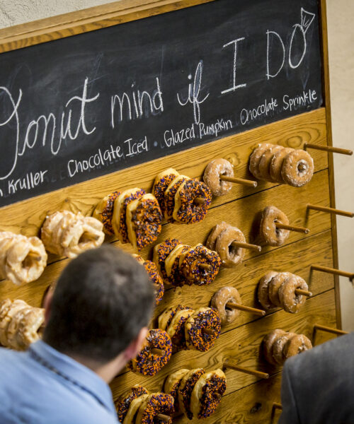 Two people stand in front of a donut wall with various donuts and a sign that reads "Donut mind if I do.