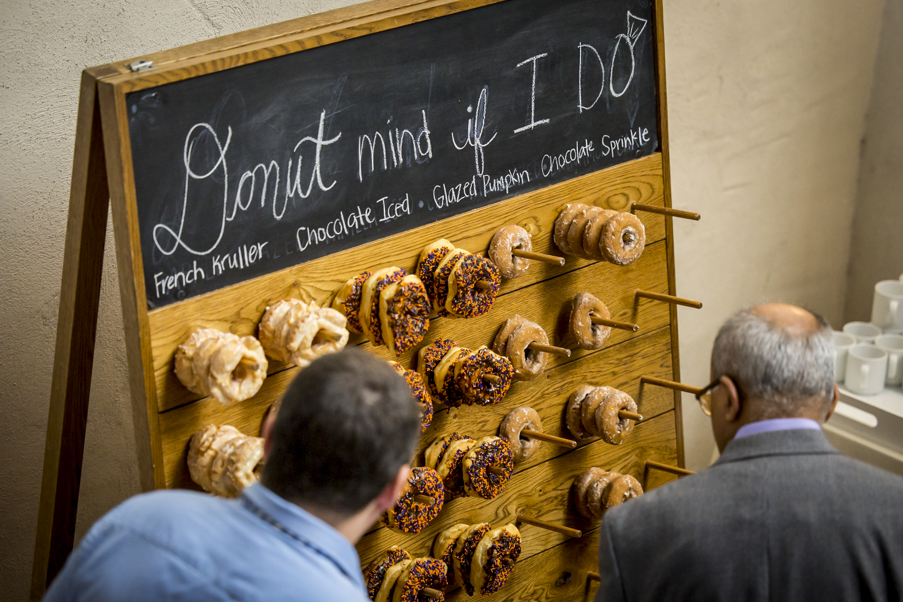 Two people stand in front of a donut wall with various donuts and a sign that reads "Donut mind if I do.